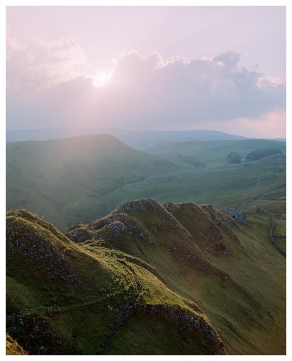 The sun shines through clouds over a valley with rolling green hills.