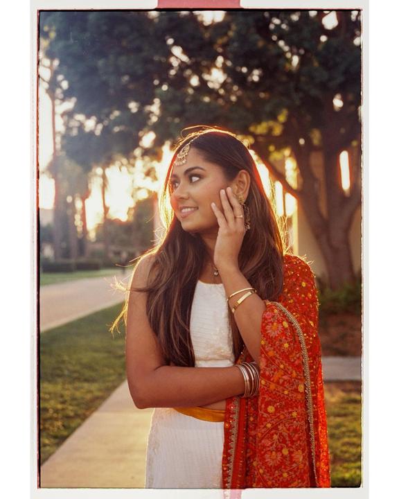 A young woman in traditional attire stands outdoors, looking to the side with a smile as the sun shines through the trees behind her.