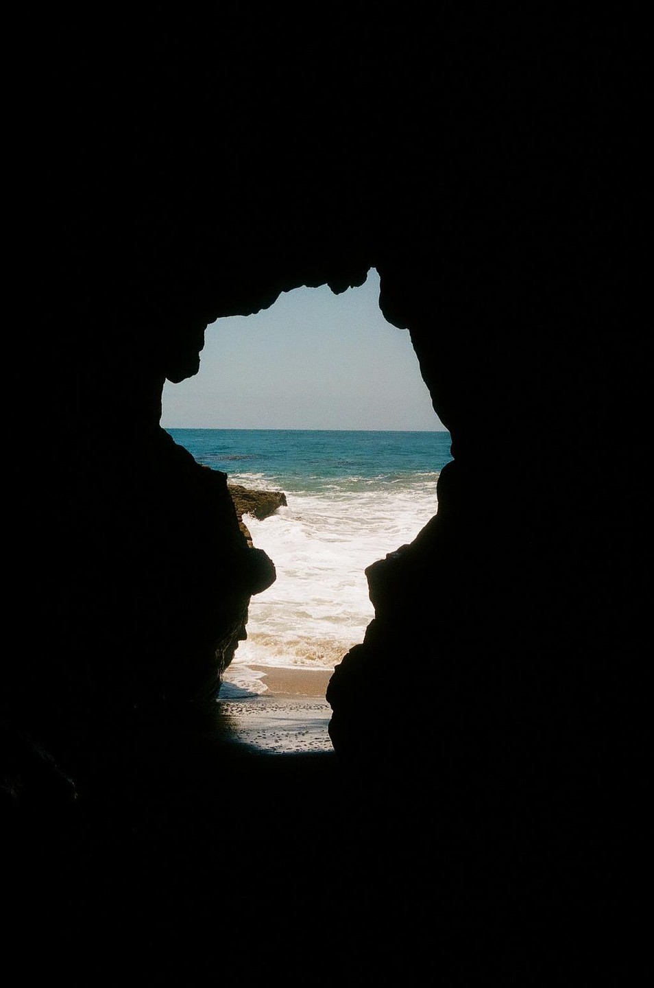 A view of the ocean and beach is framed by a dark cave opening.