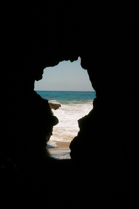 A view of the ocean and beach is framed by a dark cave opening.