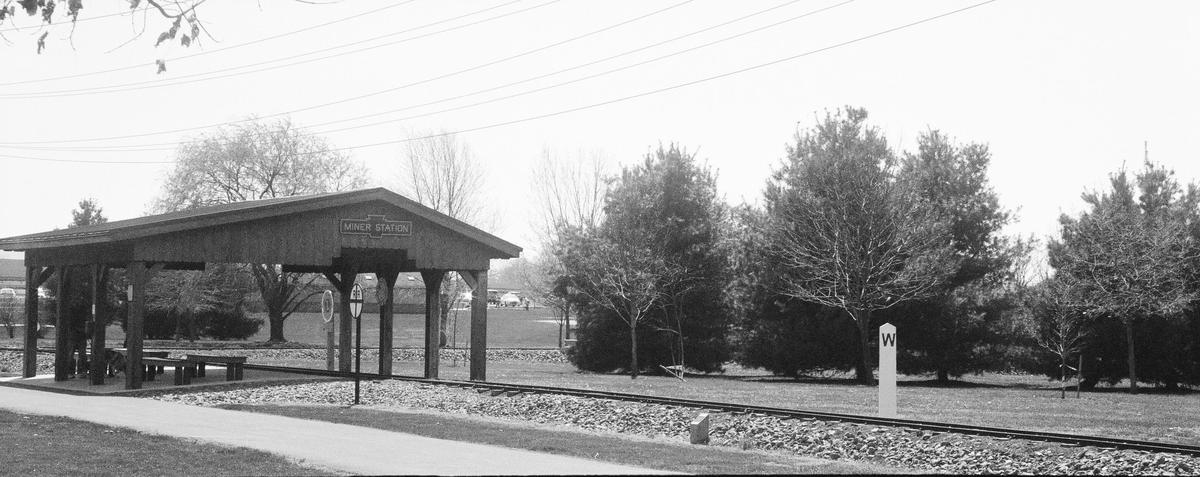 A small wooden shelter with a sign that reads "Miner Station" stands next to a railroad track, with trees and a grassy area in the background.