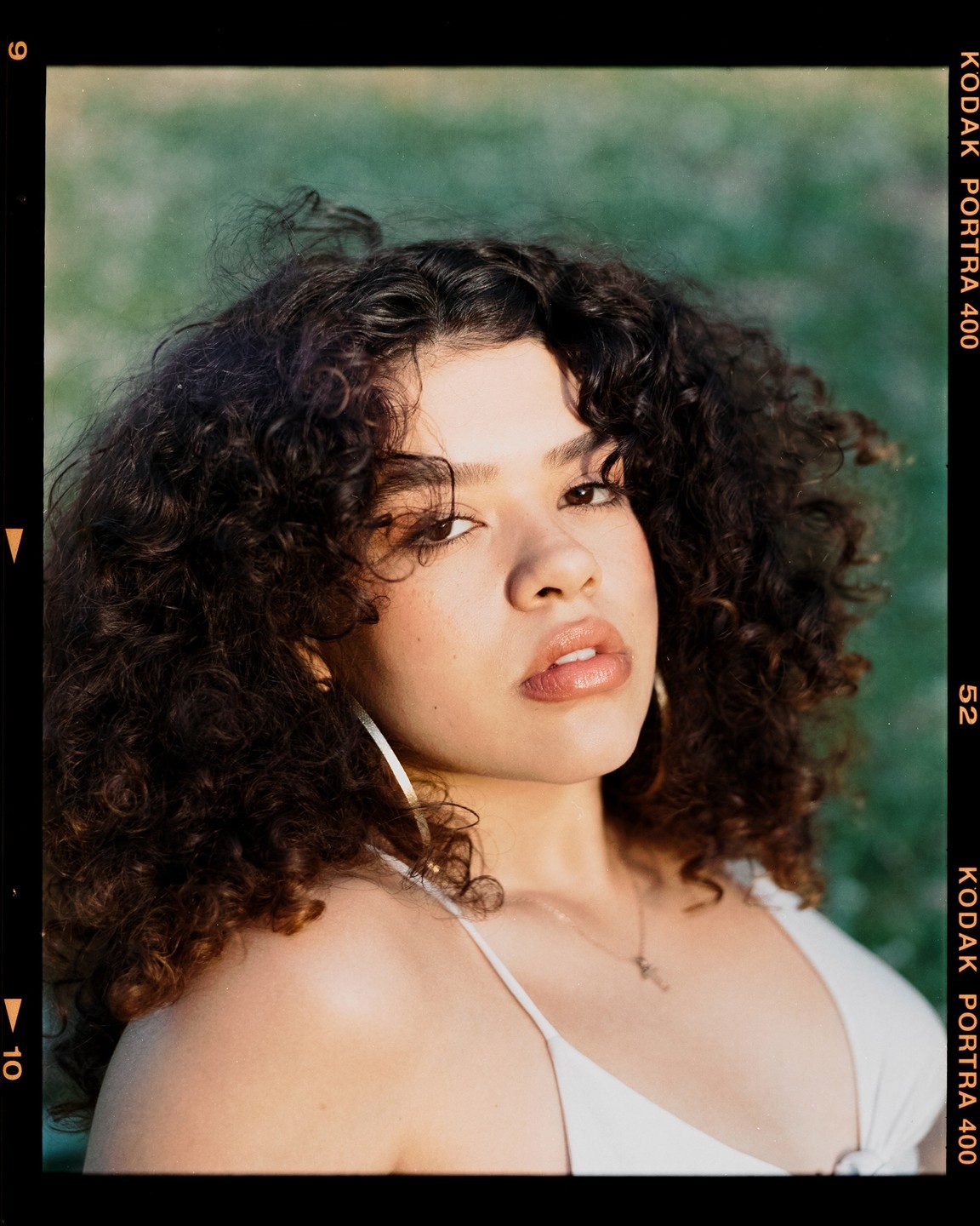 A woman with curly hair and a white top looks directly at the viewer.