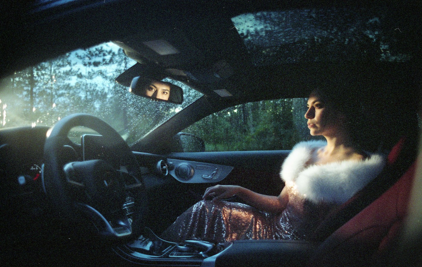 A woman in a sparkly dress and white fur stole sits in a car, looking out the window, with her eyes visible in the rearview mirror.