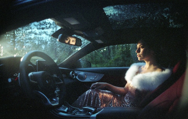 A woman in a sparkly dress and white fur stole sits in a car, looking out the window, with her eyes visible in the rearview mirror.