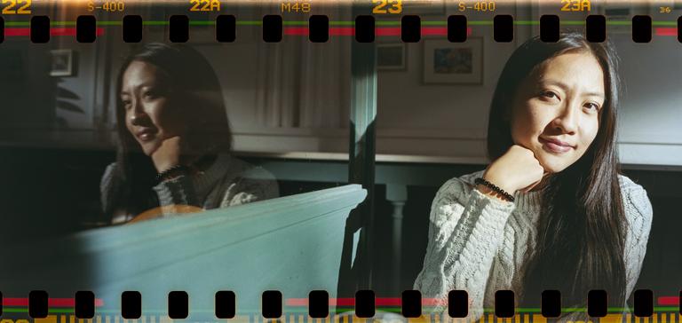 A woman with long hair and a light-colored sweater sits with her chin resting on her hand, with her reflection visible in a surface to her left.