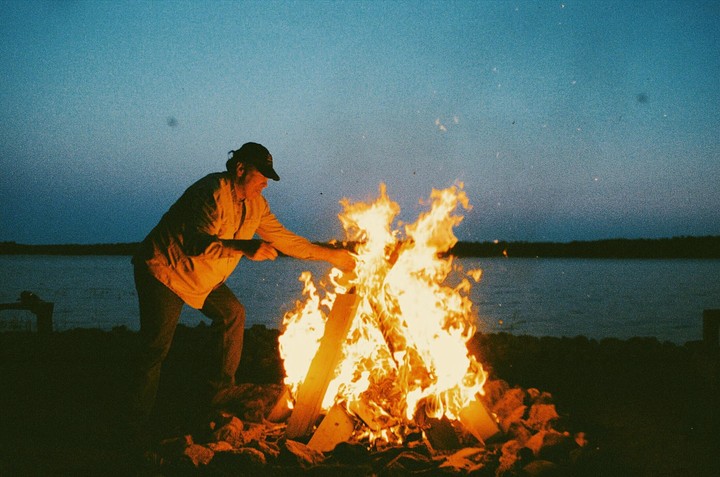 A man tends a large bonfire on the shore of a lake at dusk.