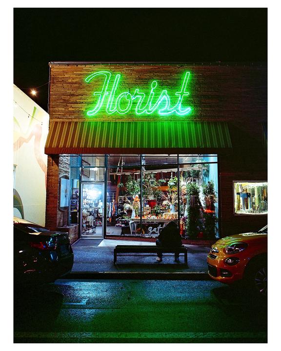 A person sits on a bench in front of a florist shop with a bright green neon sign at night.