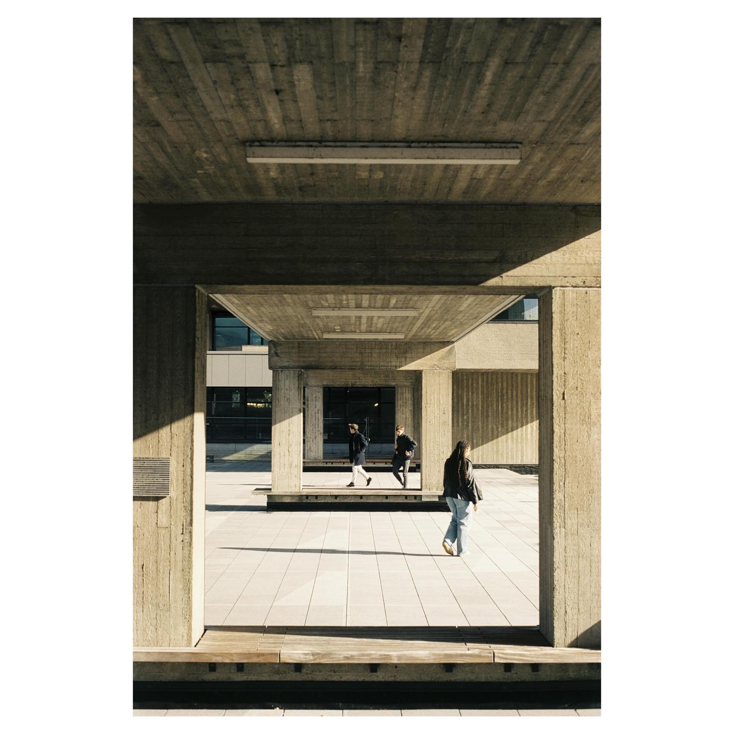 Three people walk through a concrete brutalist building with strong shadows.