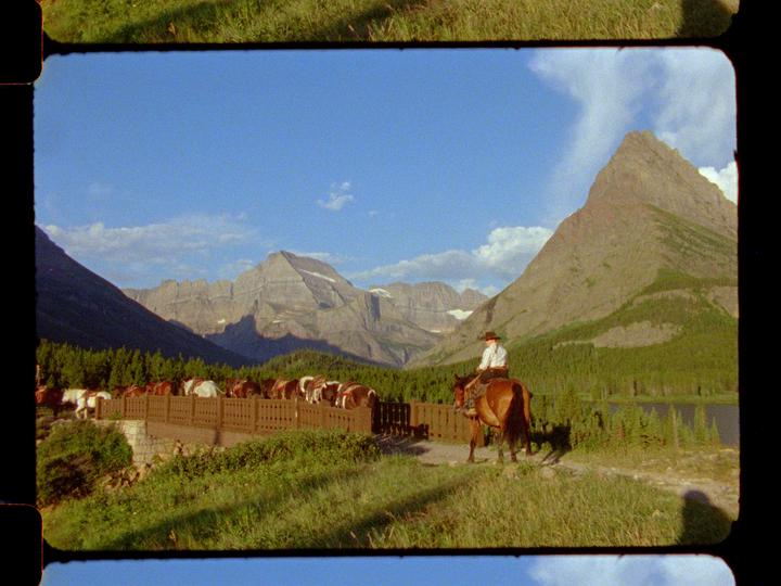 A person on horseback leads a line of horses across a bridge in a mountainous landscape.