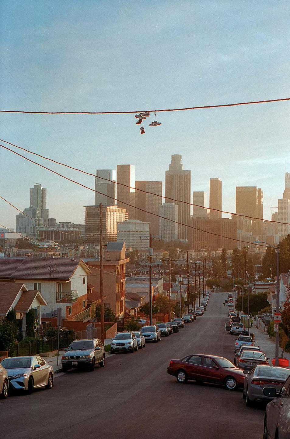 A pair of shoes hangs from a power line above a street with parked cars and a city skyline in the background.
