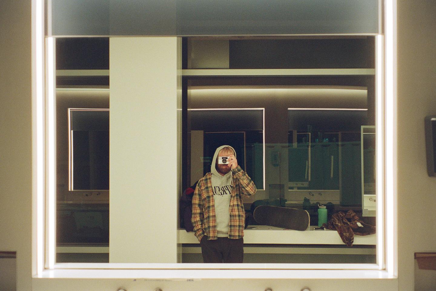 A man wearing a plaid shirt and a hoodie takes a self-portrait in a mirror.