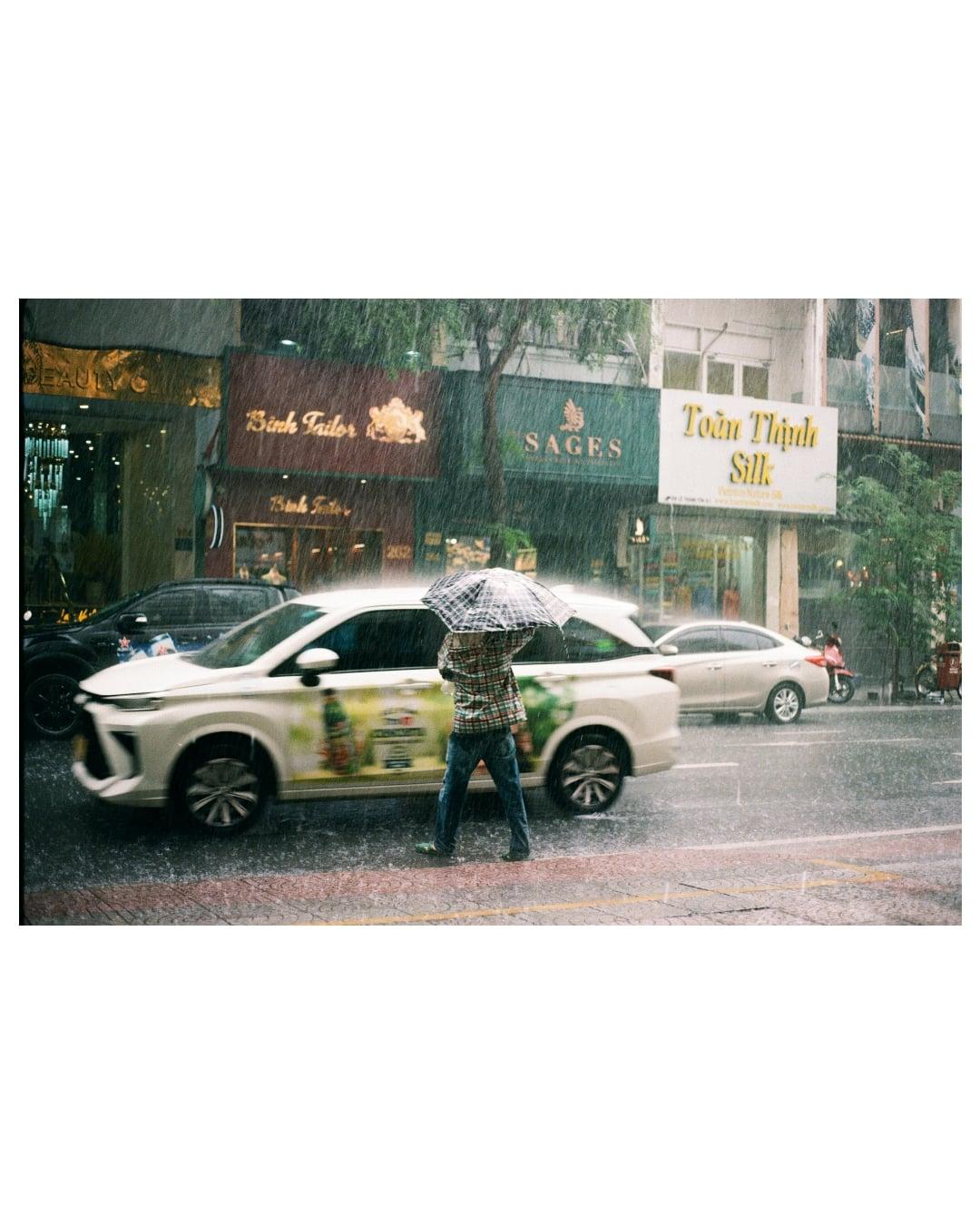 A man with an umbrella walks past a white car on a rainy street with storefronts in the background.