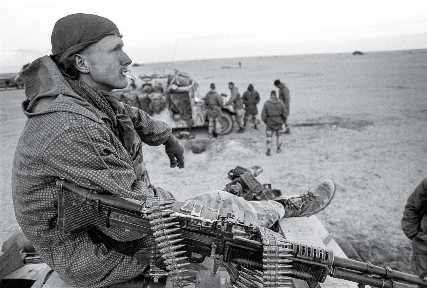 A man in military fatigues with a cigarette in his mouth sits next to a machine gun with a belt of ammunition, looking out over a desert landscape where other soldiers are gathered.
