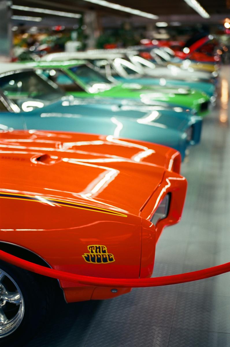 A row of vintage cars in various colors is parked indoors, with the front of an orange car in the foreground.