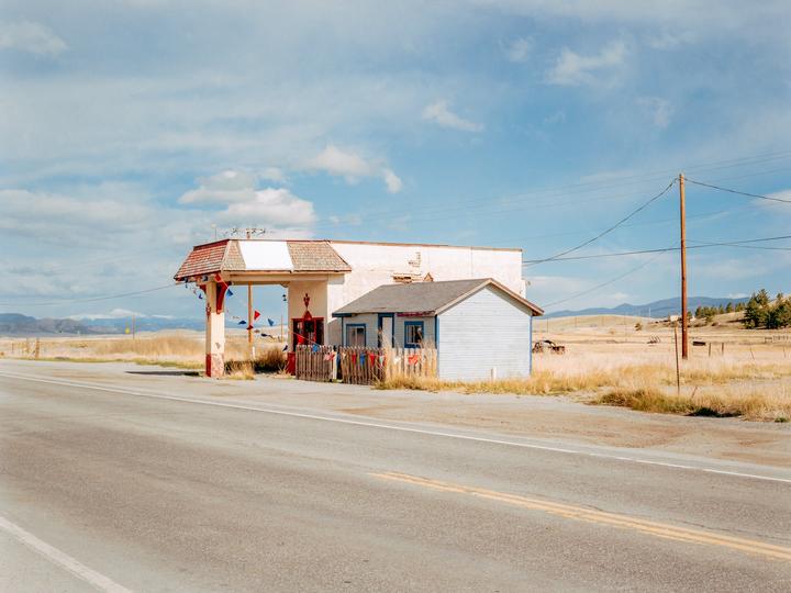 An old gas station with a small blue building next to it stands by a road in a dry, grassy landscape under a partly cloudy sky.
