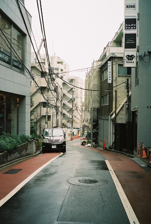 A black van is parked on a narrow street with buildings and power lines overhead.