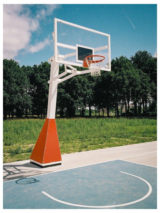 A basketball hoop stands on a blue court next to a grassy field and trees under a blue sky.