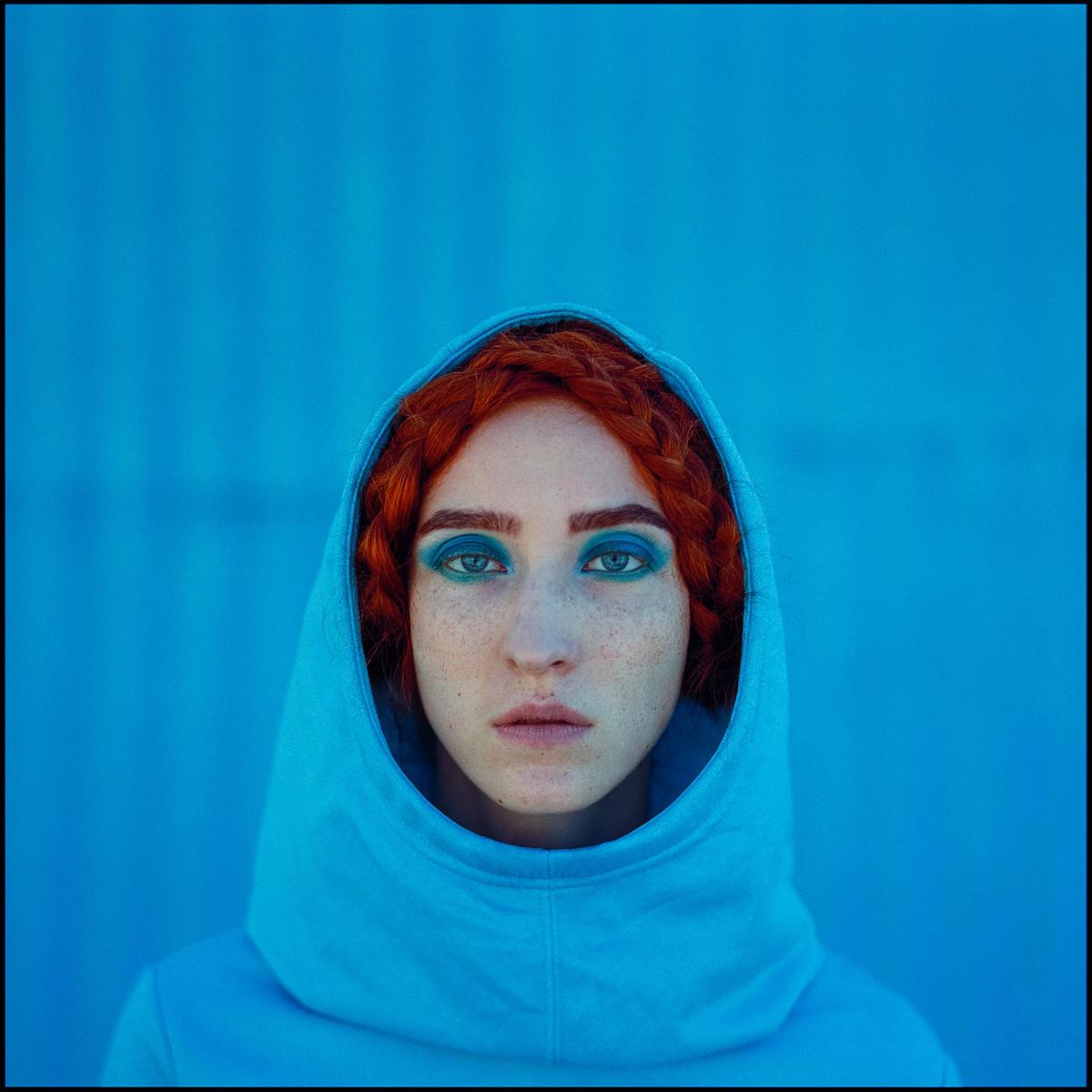 A woman with red braided hair and blue eye makeup looks directly at the viewer while wearing a blue hooded sweatshirt against a blue background.