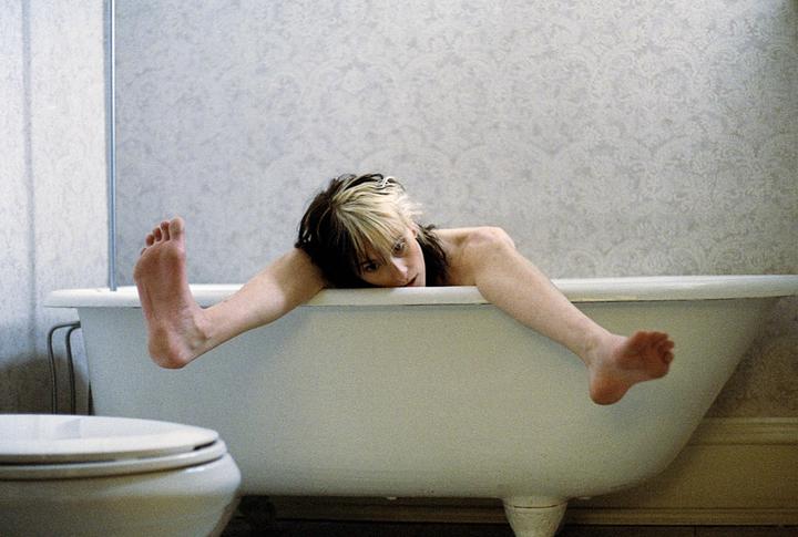 A woman with two-tone hair lies across a bathtub with her arms and legs draped over the sides.
