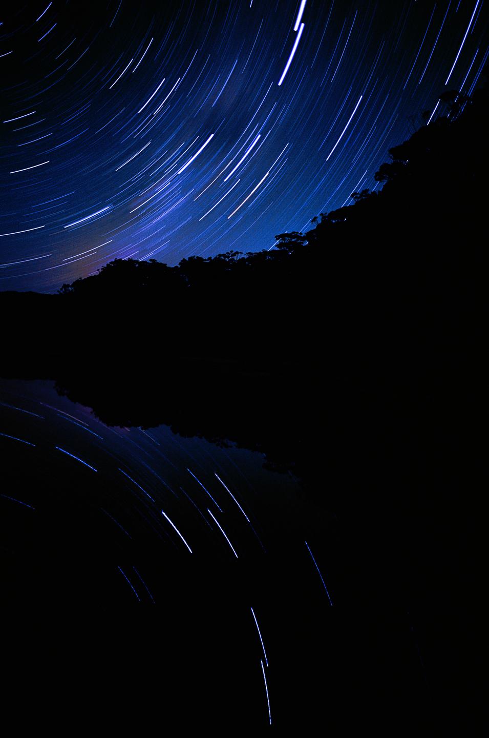 Star trails arc across a dark night sky above a silhouetted forest and its reflection in water.