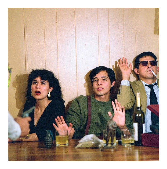 A woman and two men sit at a table with their hands raised, while a person points a gun at them.