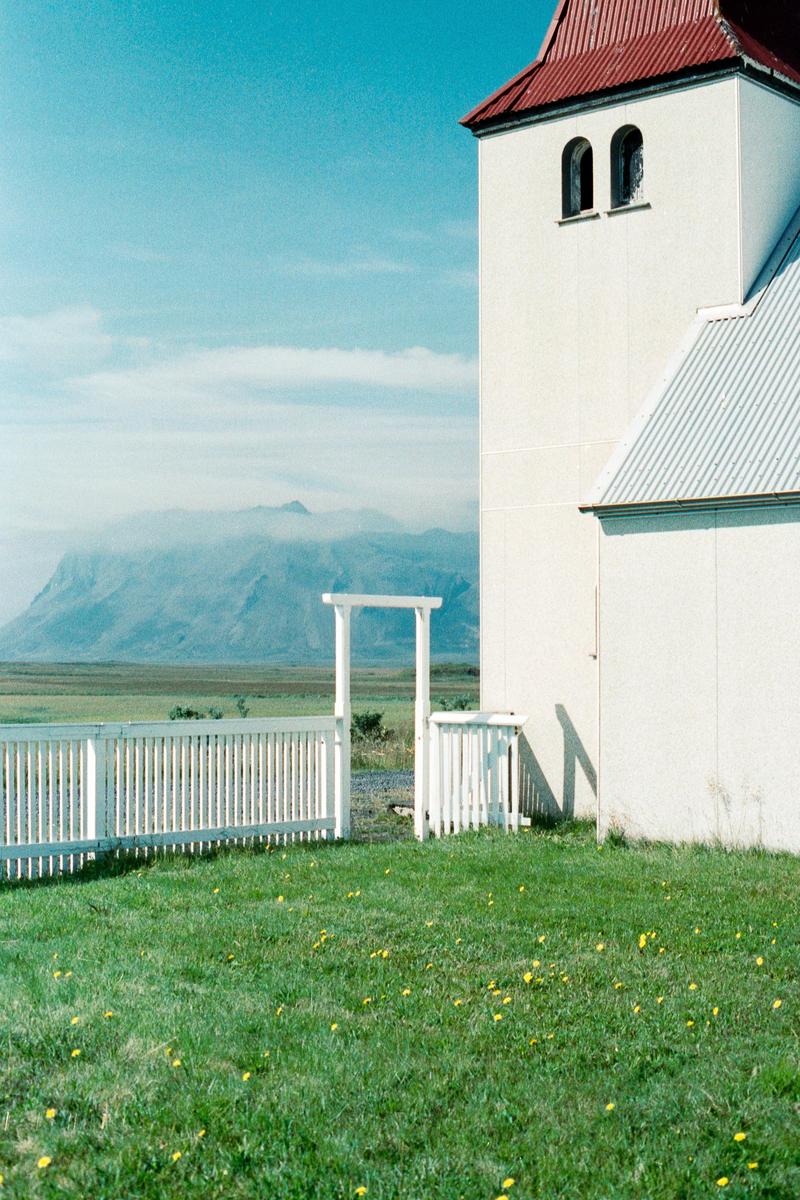 A white church with a red roof stands next to a white picket fence, with a grassy field in the foreground and mountains in the background.