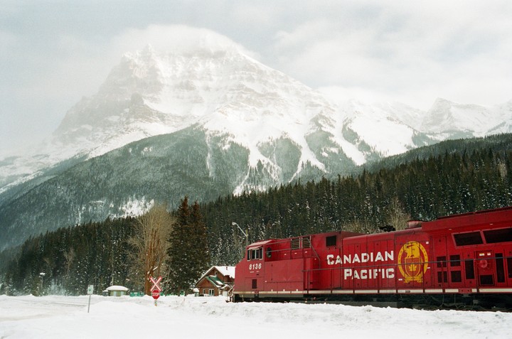 A red Canadian Pacific train is parked in a snowy landscape with a dense forest and a large, snow-covered mountain in the background.