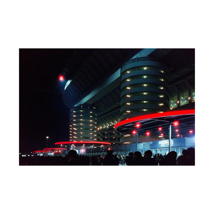 A crowd of people gathers outside a large stadium with red and white lights at night.