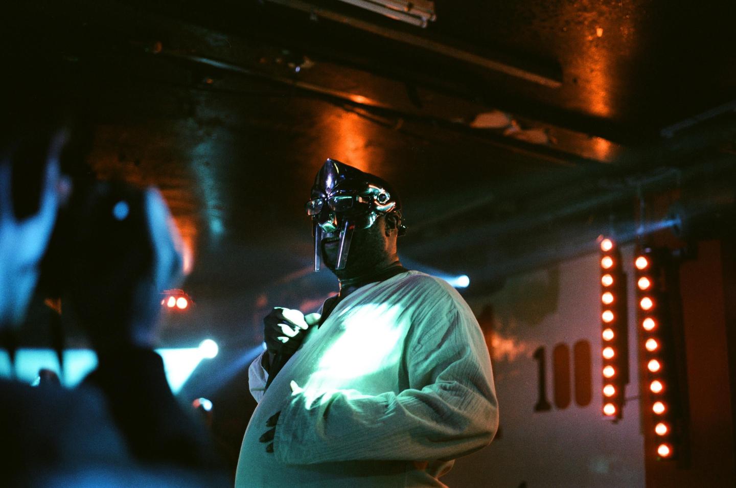 A man wearing a metal mask and a light-colored shirt stands on a stage with colorful lights.