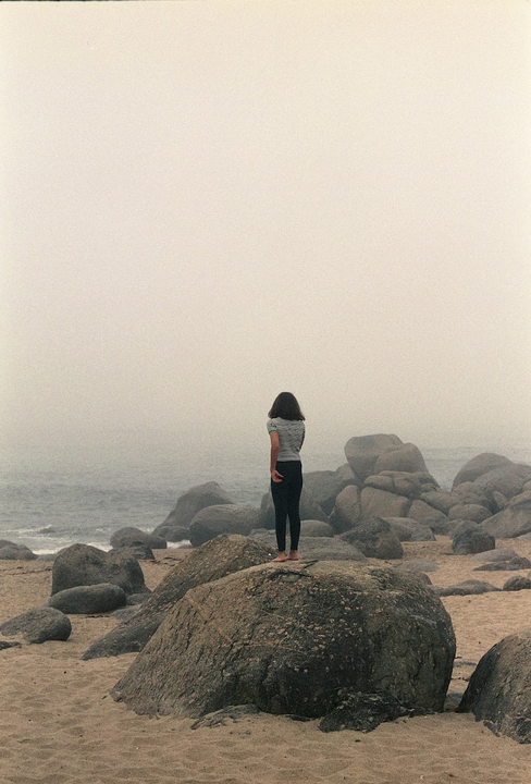 A woman stands barefoot on a large rock on a foggy beach with many boulders.