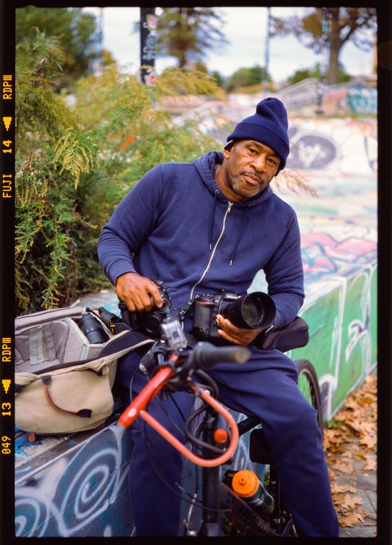 A man sits on a bicycle holding two cameras, with a camera bag next to him and a graffiti-covered wall in the background.