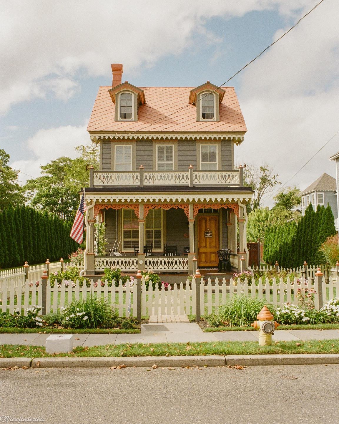A charming Victorian house with a pink roof and a white picket fence stands under a partly cloudy sky.