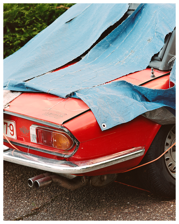 A red car is partially covered by a torn blue tarp.