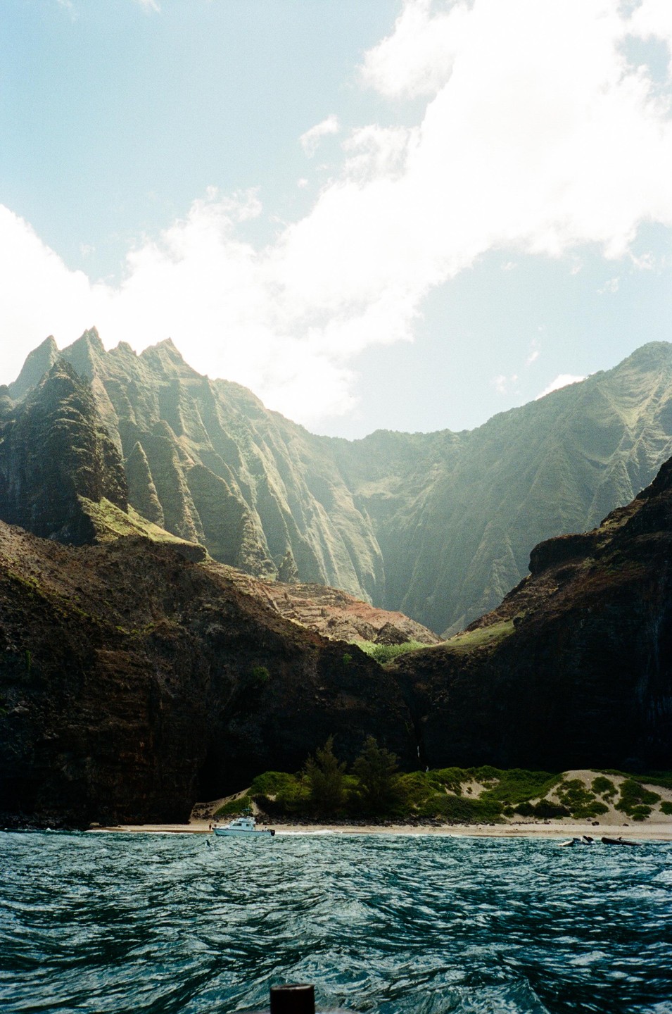 A boat is beached on a small sandy shore at the base of large, steep mountains that rise from the ocean.