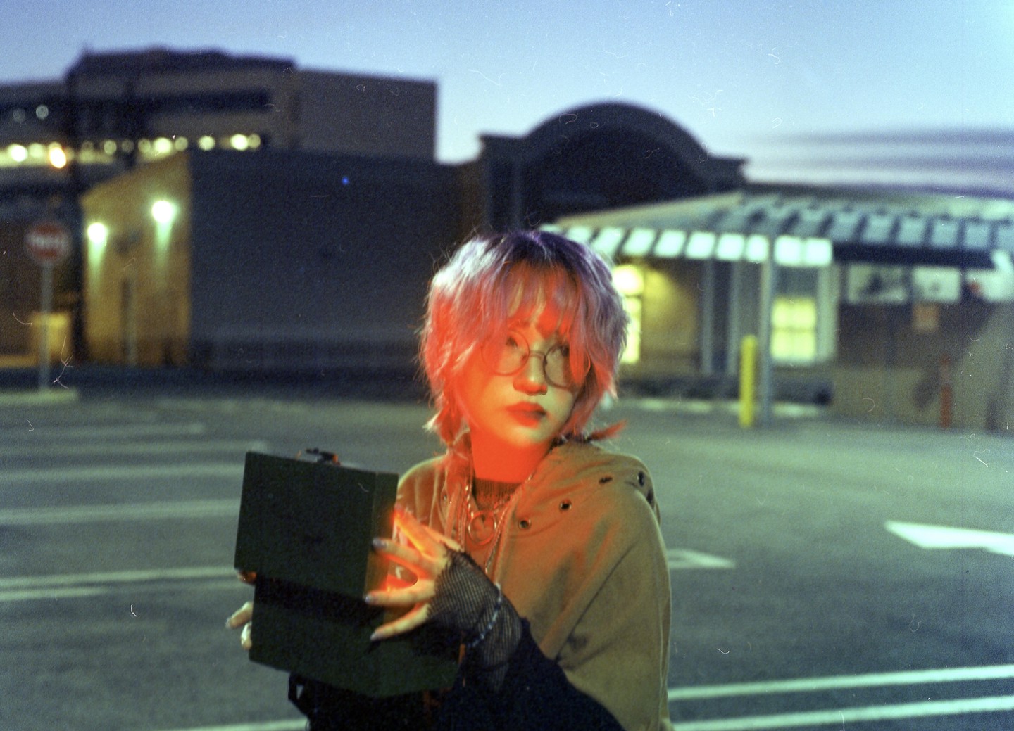 A woman with pink hair and glasses holds an open box with glowing light in a parking lot at night.