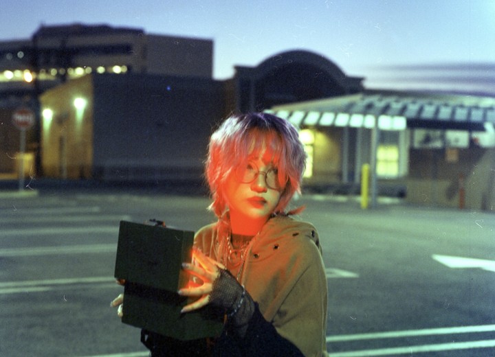 A woman with pink hair and glasses holds an open box with glowing light in a parking lot at night.