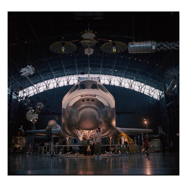 A space shuttle is displayed in a large hangar with a woman standing in front of it.