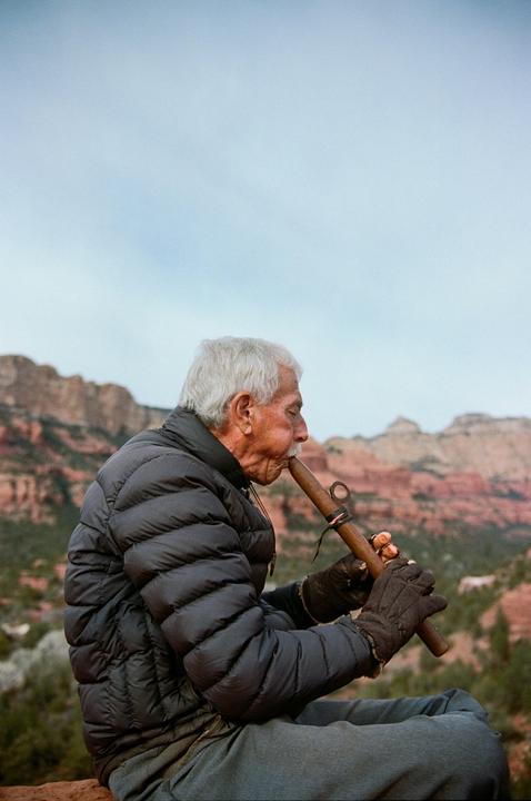A man plays a wooden flute with red rock mountains in the background.