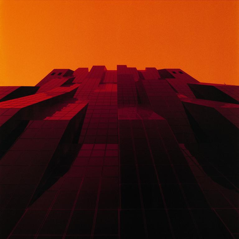 A large, dark, geometric building with many windows is visible against an orange sky.