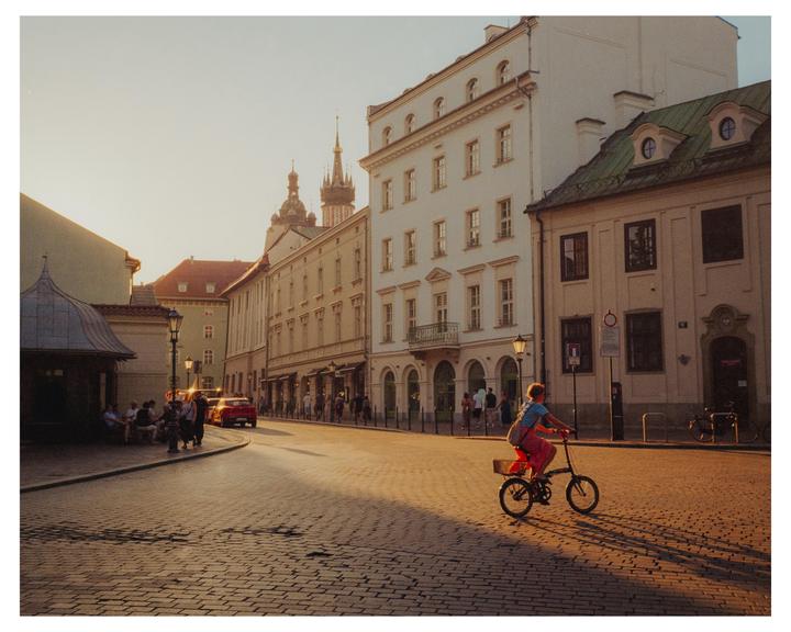 A woman rides a bicycle down a cobblestone street lined with buildings and people during golden hour.