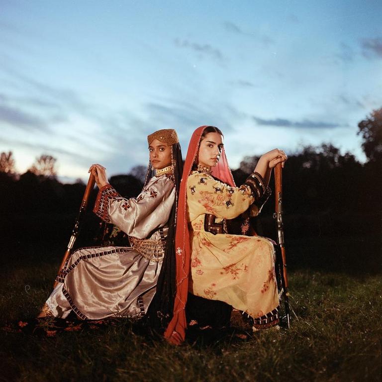 Two women in traditional clothing sit back-to-back in a field at dusk, each holding a rifle.