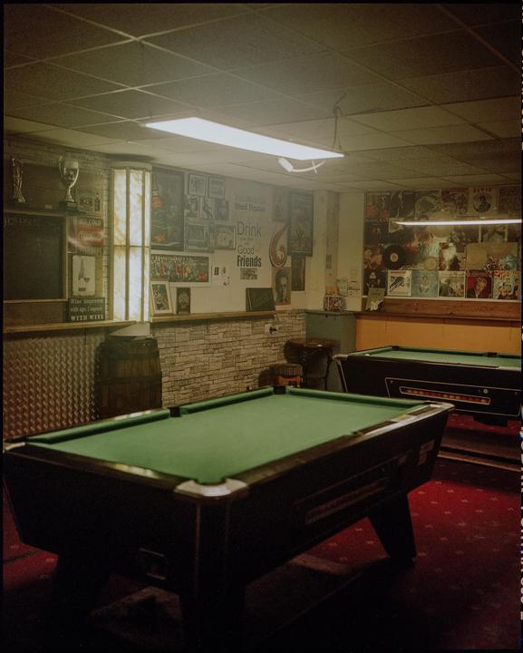 Two pool tables with green felt stand on a red carpet in a room with posters and signs on the walls.