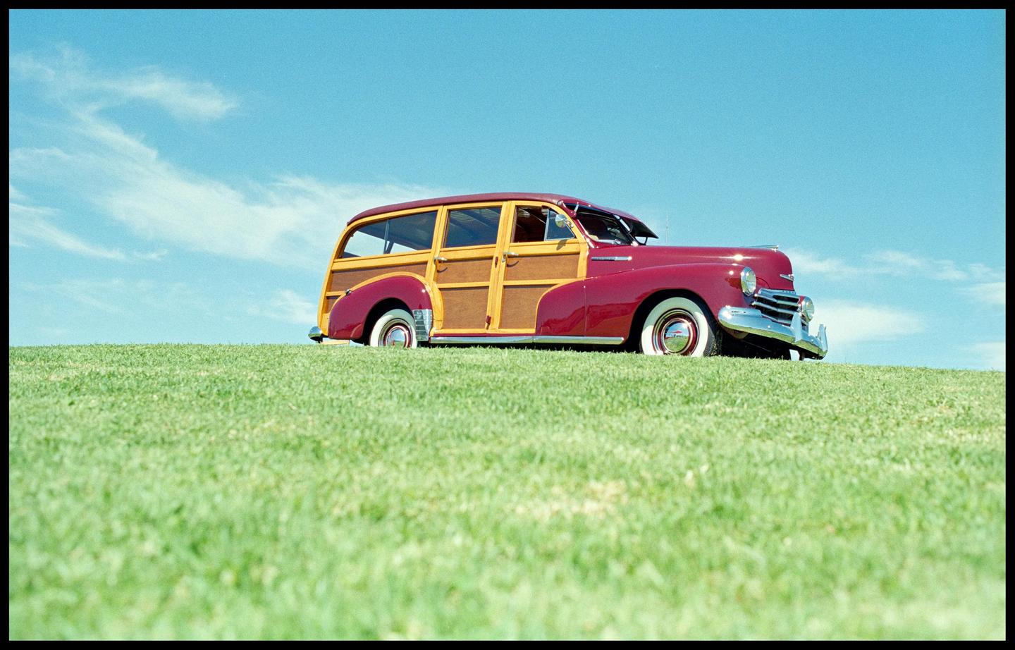 A red and wood paneled station wagon is parked on a grassy hill under a blue sky.