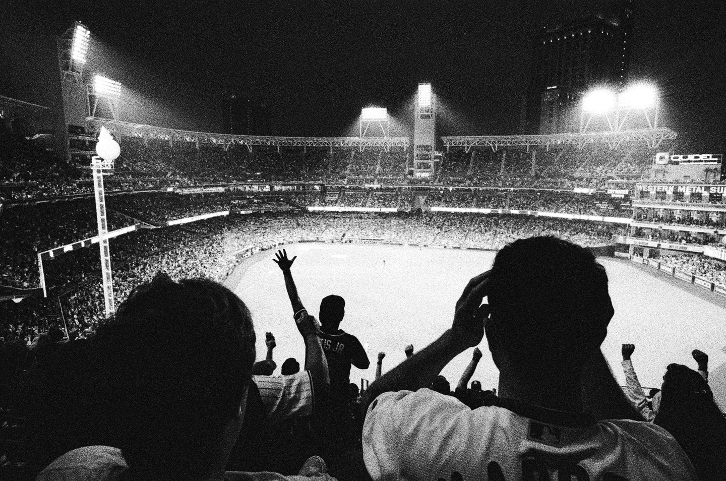 A baseball stadium is illuminated by bright lights at night, with spectators in the foreground watching the game.