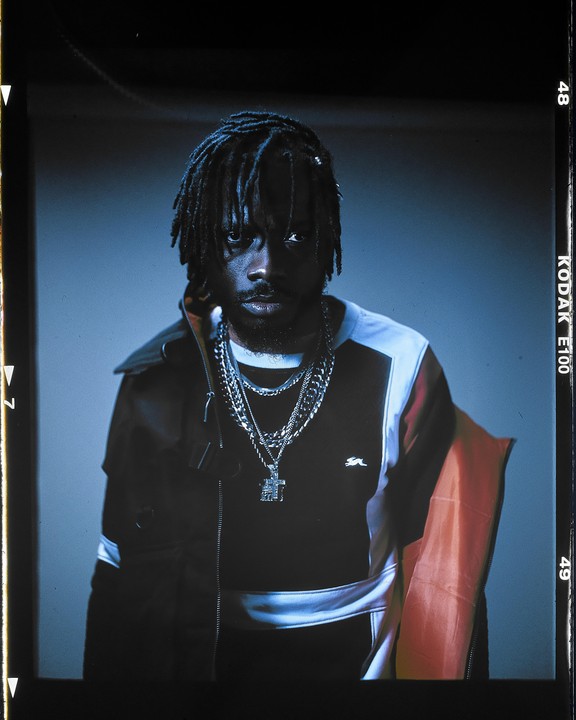 A man with dreadlocks and multiple chain necklaces looks directly at the viewer.