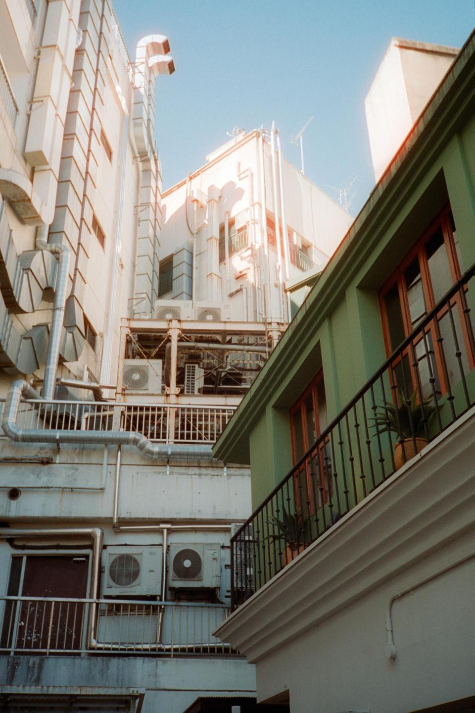 A narrow alleyway is visible between two buildings, one with a green facade and a balcony, and the other with many pipes and air conditioning units.