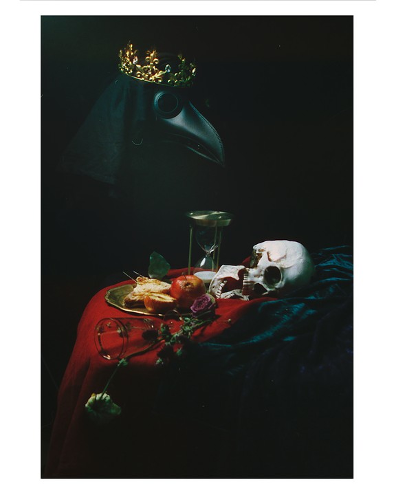 A plague doctor mask with a crown, a skull, an hourglass, and fruit are arranged on a red and blue draped table.