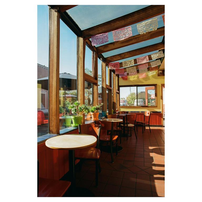A bagel shop interior is filled with tables and chairs, with plants on a window sill and colorful decorations hanging from the ceiling.