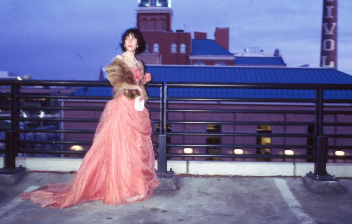 A woman in a long pink gown and fur stole stands on a rooftop at dusk.