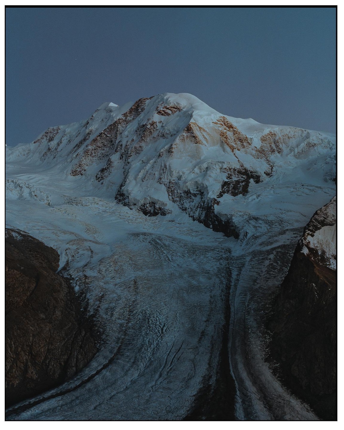 A snow-covered mountain peak rises above a glacier at dusk.